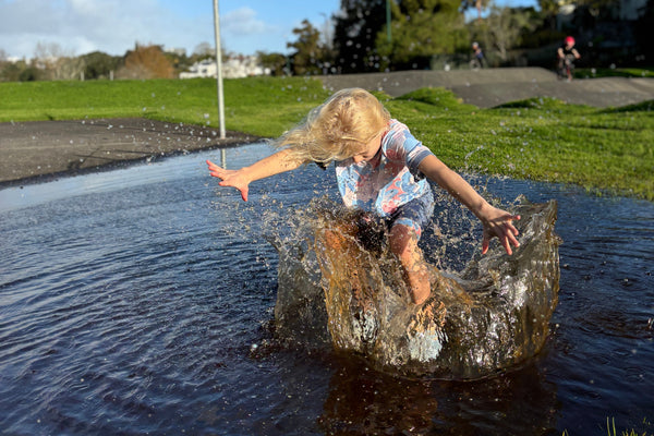Child standing in a puddle with a clear blue sky and green grass in the background wearing aszo blue and pink tee and shorts. sensory-friendly clothing.