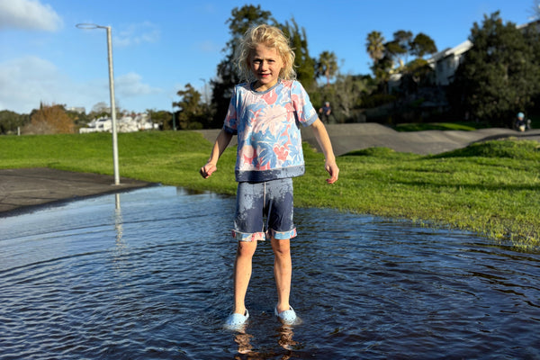 Child standing in a puddle with a clear blue sky and green grass in the background wearing aszo blue and pink tee and shorts. sensory-friendly clothing.