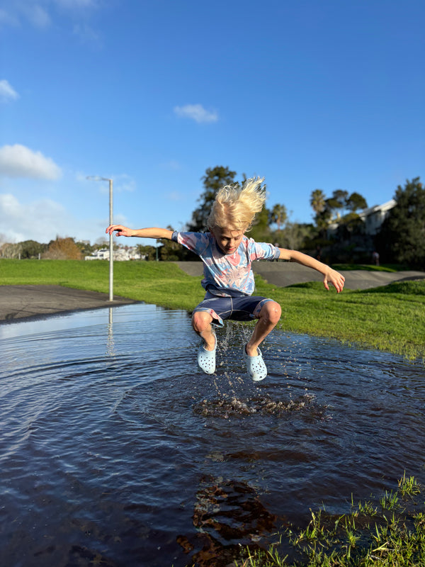 Child standing in a puddle with a clear blue sky and green grass in the background wearing aszo blue and pink tee and shorts. sensory-friendly clothing.