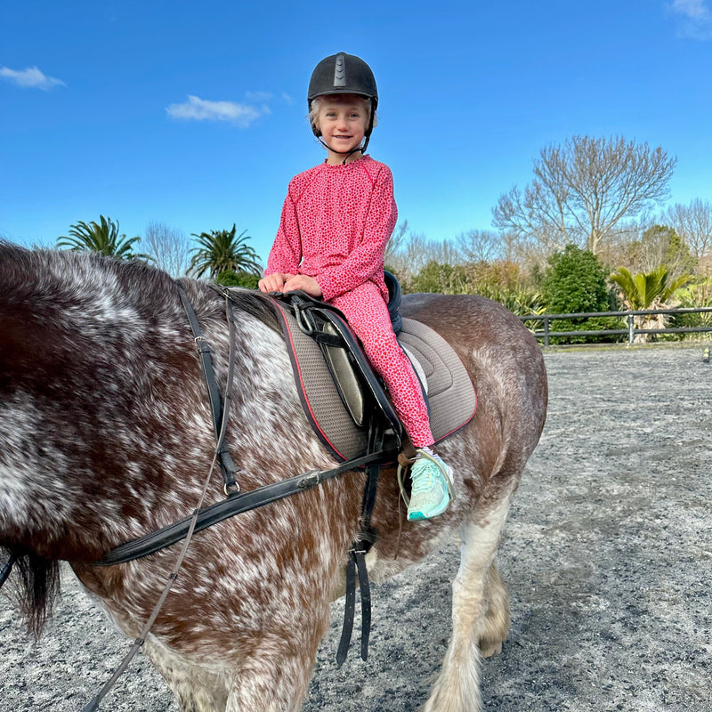 Child  wearing aszo apparel, sensory-friendly top and pants,  in pink outfit riding a horse on a clear day. clothing aszo.